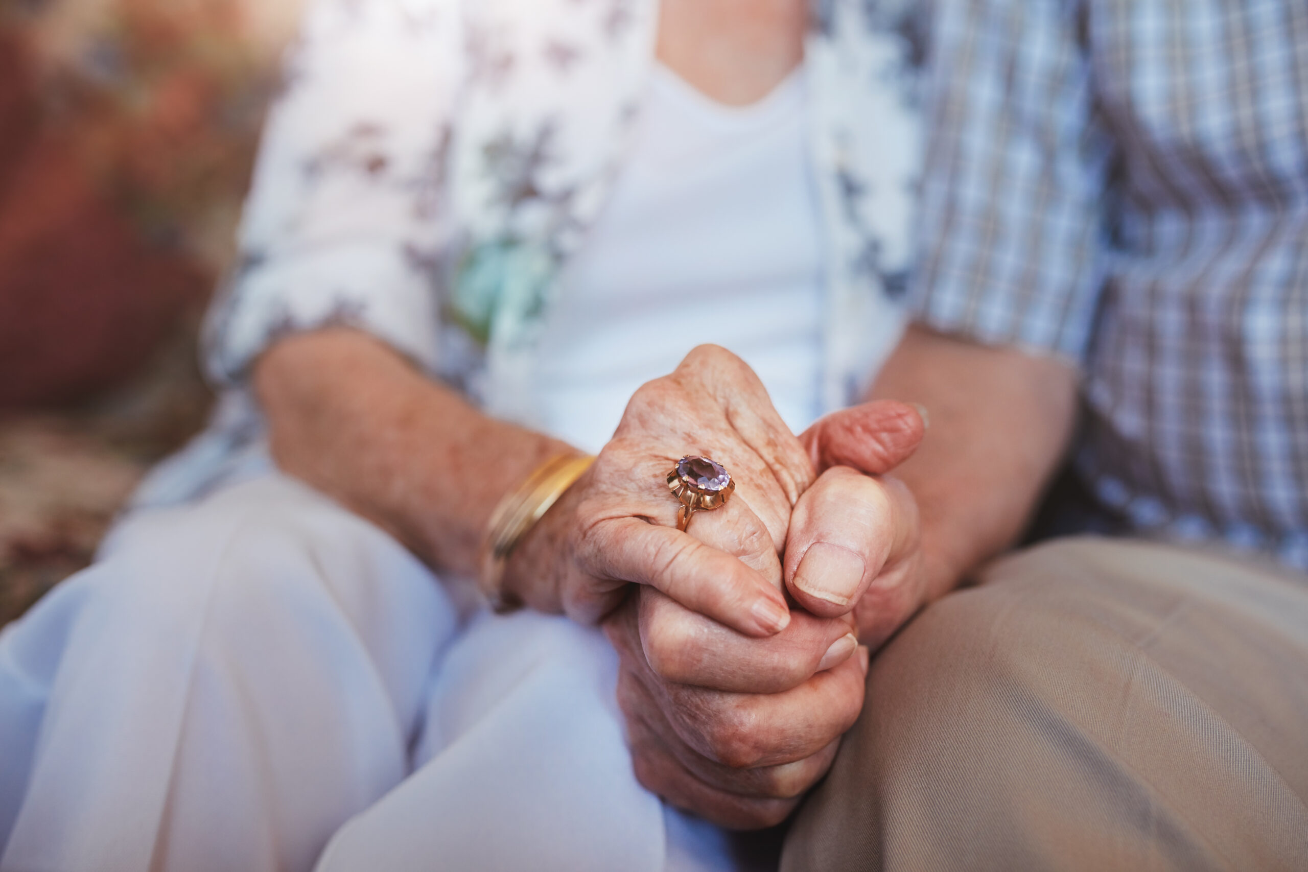 Cropped shot of elderly couple holding hands while sitting together at home - Care Partners
